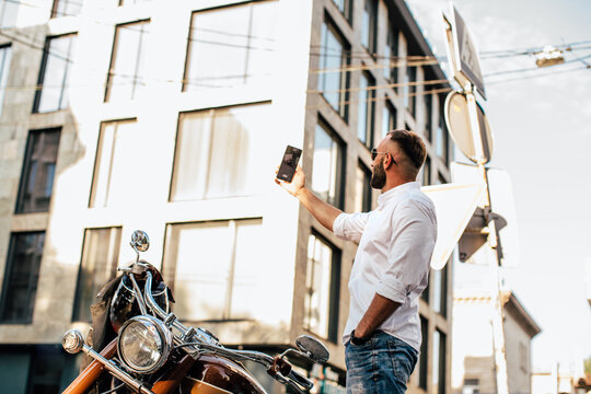 Elegant man taking selfie with smartphone near motorcycle in city