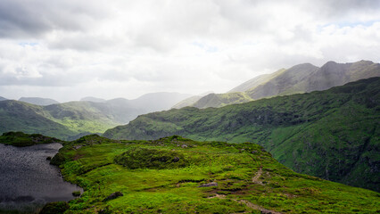 A scenic vista shows layers of rolling green hills and a tranquil lake from an overlook in the Gap of Dunloe, County Kerry. Overcast sky with scattered light accentuates the landscape.