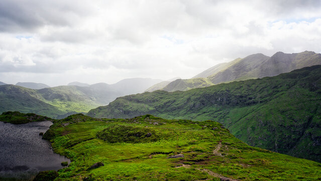 A scenic vista shows layers of rolling green hills and a tranquil lake from an overlook in the Gap of Dunloe, County Kerry. Overcast sky with scattered light accentuates the landscape.