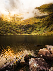 This showcases the still waters of Lough Callee, reflecting the surrounding hills. A small waterfall cascades down the slope into the calm lake, nestled in the Irish countryside.