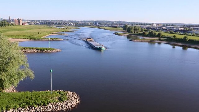 Een lang schip vaart over de Nederrijn/ rivier de Rijn, vlakbij de stad Arnhem. De stad zie je nog op de achtergrond. De groene uitwaarden zijn zichtbaar en het water van de rivier staat redelijk laag