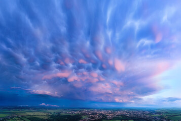 Panoramic view of vibrant mammatus clouds at sunset over a city and green fields. Ideal for: weather background, dramatic skies, nature prints, and meteorological visual content.