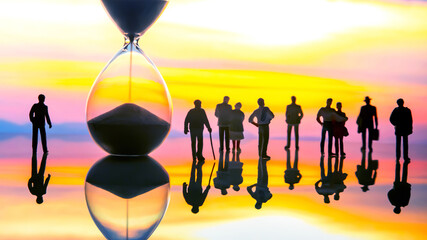 Silhouettes of people stand before a giant hourglass at sunset, reflecting on a glossy surface,...
