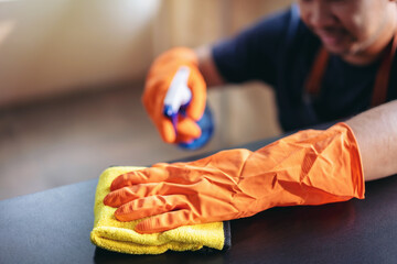 Housekeeper man in orange rubber gloves using a clean spray and a duster while cleaning in living room at home, housekeeping and cleaning concept