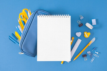 School background with empty notebook in the center of the composition and full pencil case with stationery and school supplies including sharpener, paper clips on blue flat background.