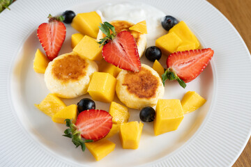Cottage cheese pancakes are lying on a white plate with sour cream or yogurt, decorated with fresh mango, strawberries and blueberries