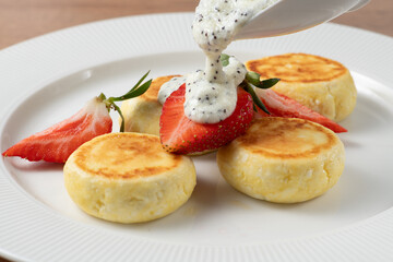 Chef is pouring poppy seed sauce on a plate of syrniki decorated with fresh strawberries, a traditional Eastern European dish