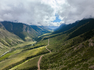 A high-angle aerial view of a winding dirt road traversing a vibrant green mountain valley, with a...
