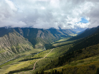 Fototapeta premium A sweeping aerial view captures a vast mountain valley with a river winding through its base, a dirt road on the slopes, and dramatic clouds overhead