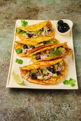 Vegetable tacos with zucchini, carrot and potato tortilla filled with canned tuna, sliced ​​avocado and olives on ceramic plate on green concrete background.