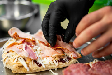 A chef wearing black gloves prepares a fresh sandwich with bacon, cheese, and sauce in a professional kitchen. The close-up captures the detailed cooking process and presentation