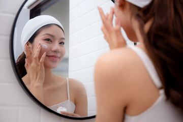 Beautiful young asian woman applying cream or lotion for rejuvenation skin on face while looking mirror in bathroom, woman applying moisturizer for treatment wrinkles on facial, beauty and skincare.