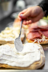 A close-up of a hand spreading creamy white sauce on a flatbread with a spoon. Other flatbreads with various toppings like cheese and sliced meats are visible in the background. preparation process.