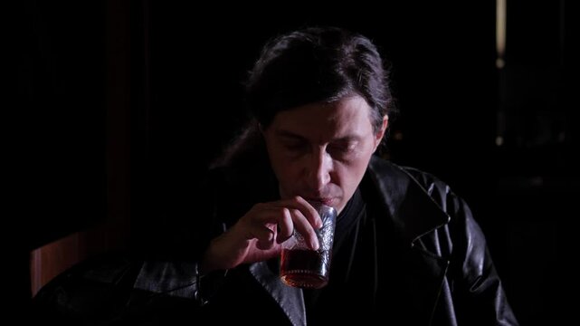 Close-up shot captures a somber and introspective mood as a man in a dark leather jacket sips from a glass, eyes closed, against a deep black background, suggesting a moment of solitude or reflection