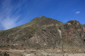 View Of A Barren Rocky Mountain Slope With Several Power Line Towers Against A Deep Blue Sky With Wispy Clouds.