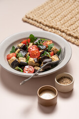 Eggplants tomatoes arugula salad in gray bowl on light beige background. Vegetarian summer Mediterranean salad