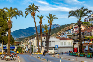 Fethiye square near marina with tall date palms, taxis and scooters against mountain backdrop under evening sun. Vibrant Mediterranean urban scene. Fethiye, Turkey.