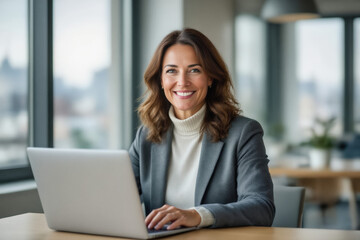 A smiling businesswoman with shoulder-length brown hair sits at a desk, looking directly at the camera while typing on a laptop.