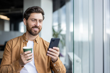 Successful male businessman inside office standing by window with phone in hands and cup of hot...