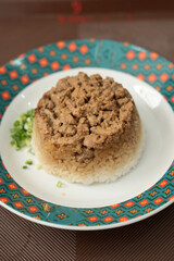 Nasi tim, white rice topped with pieces of spiced chicken, formed into a bowl, and sprinkled with chopped spring onions on a patterned plate.