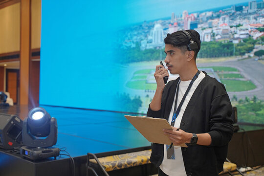 Asian male stage manager directs the show on stage using a headset, walkie-talkie, and clipboard, showing the professional work atmosphere behind the scenes of a show production.