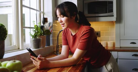 Asian woman reading smartphone messages, placing voice call, gazing outside in kitchen - Powered by Adobe