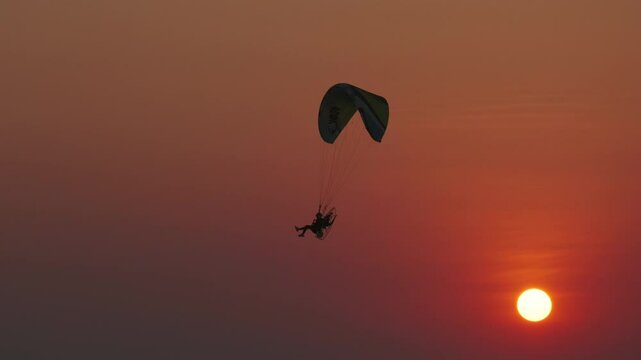Motor paraglider flying through colorful sunset sky and setting orange sun, 4k