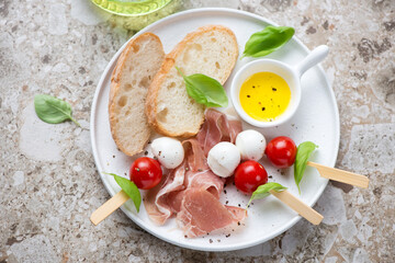 White plate with ciabatta, prosciutto and caprese skewers, horizontal shot on a light-brown granite background, elevated view