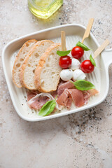 Serving tray with torn prosciutto, caprese skewers and ciabatta, vertical shot on a beige granite background, elevated view