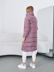 Model wearing a long, purple puffer coat, stands in front of a mirror, with their back turned. Cozy...