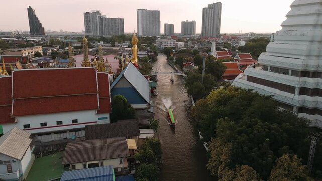 Aerial view of a longtail boat cruising along a bangkok canal, passing by wat paknam phasi charoen temple at sunset, Thailand.