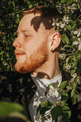 A young red-haired man with a beard in a blooming apple orchard, close-up.