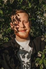 A young red-haired man with a beard in a blooming apple orchard, close-up.