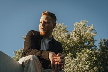 Young red-haired bearded man in a blooming apple orchard on a summer day in the park.