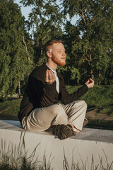 A young red-haired man with a beard sits in the lotus position and meditates in a blooming apple orchard.