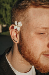 Close-up of a young red-haired bearded man with a white flower behind his ear on a summer day in the park.