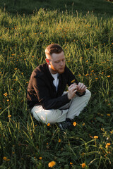 A young red-haired man with a beard sits on a blooming meadow at sunset.