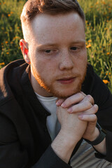 A young red-haired man with a beard sits on a blooming meadow at sunset,close-up.