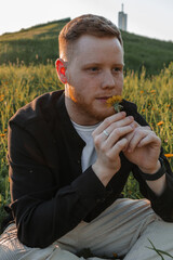 A young red-haired man with a beard sits on a blooming meadow at sunset.A man holds a yellow dandelion and inhales the scent of flower,close up.