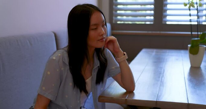 Asian woman gazing at window with orchid then pushing back and leaning on kitchen island smiling