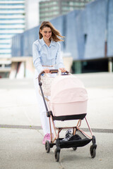 Full body smiling young woman walking through a modern cityscape while pushing baby stroller