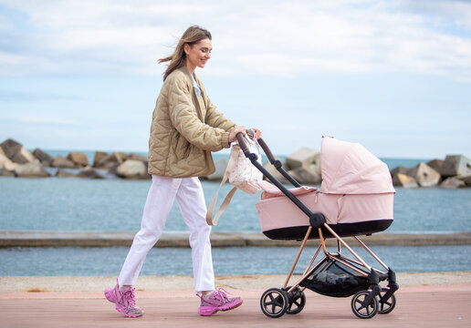 Full body young mother walks peacefully along a seaside promenade, pushing a pink baby stroller with a smile