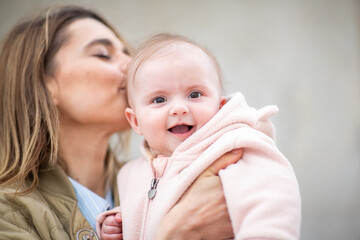 smiling baby in a pink fleece is held by her mother, who kisses her gently on the head