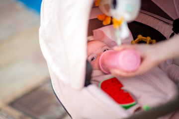 baby lies comfortably in a stroller while being fed from a pink bottle