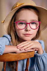 An attractive young girl of Slavic appearance with light-brown hair, wearing glasses with pink frames and a straw hat with a yellow ribbon, sits with her arms crossed on the back of a chair 