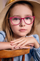 A close-up portrait of a young girl with expressive large eyes and a clear gaze. She sits with her arms crossed on the back of a chair. There is a neutral beige background behind her.