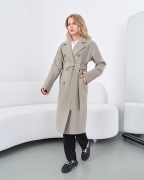 Blonde woman in a long beige trench coat with belt, standing in a modern room with a white couch and mirror, hands in pockets, wearing black shoes and black pants