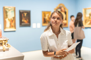 Young woman with sheet piece of paper in hands walks around museum hall, examines showpiece painting and icons at exhibition. List of recommended exhibitions, guidebook, and map of museum.