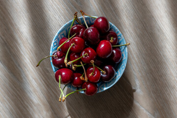  Top down view of ripe red cherries in a patterned blue bowl on a wooden surface with natural light and shadows