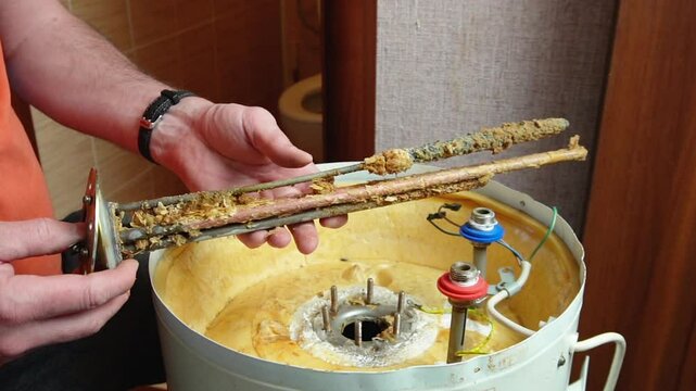 A worker holds a heating element of an electric boiler covered in rust and mineral deposits. Boiler maintenance and repair